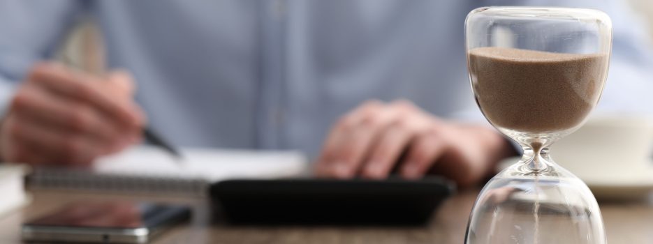 hourglass with flowing sand desk man taking notes while using calculator indoors selective focus