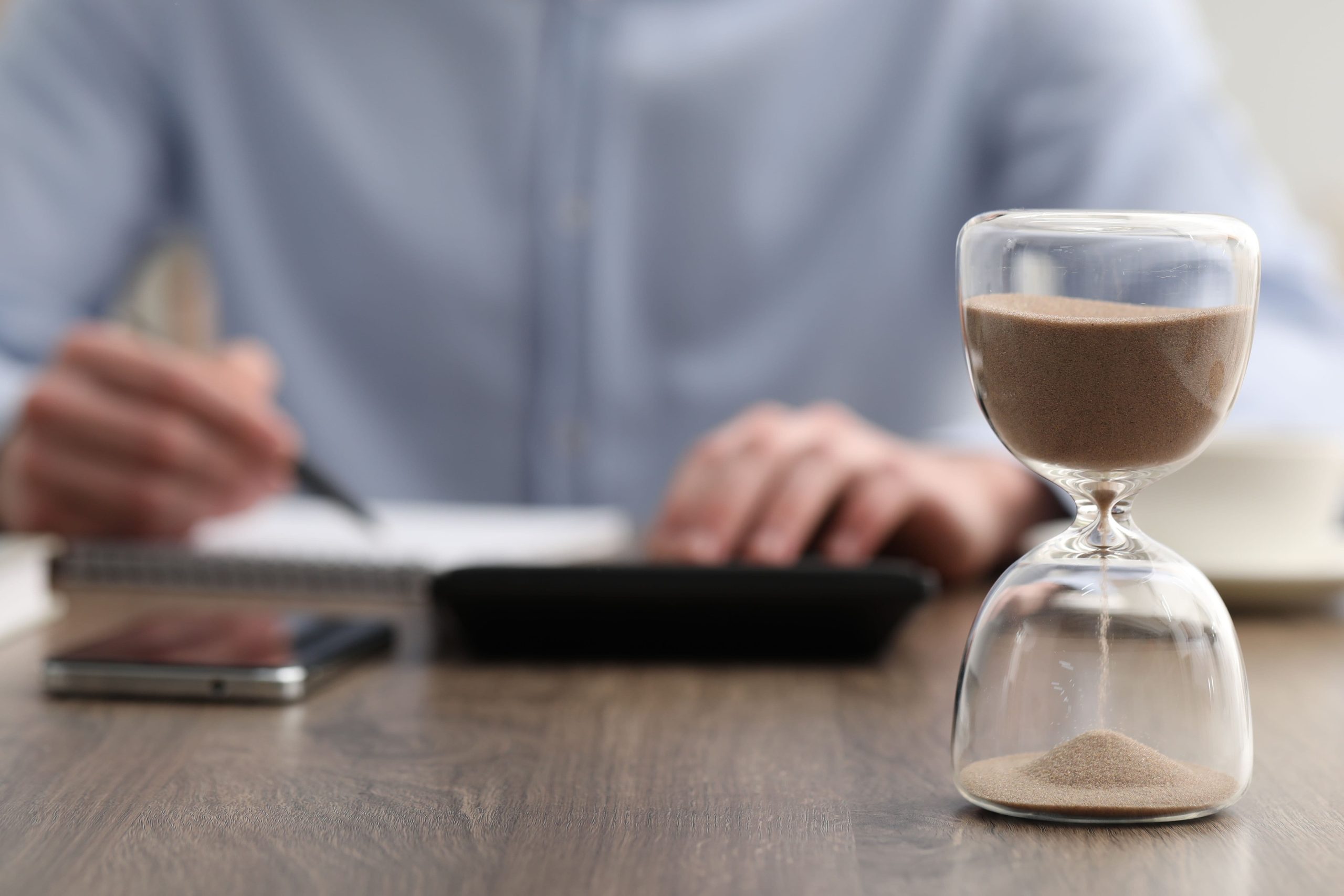 hourglass with flowing sand desk man taking notes while using calculator indoors selective focus